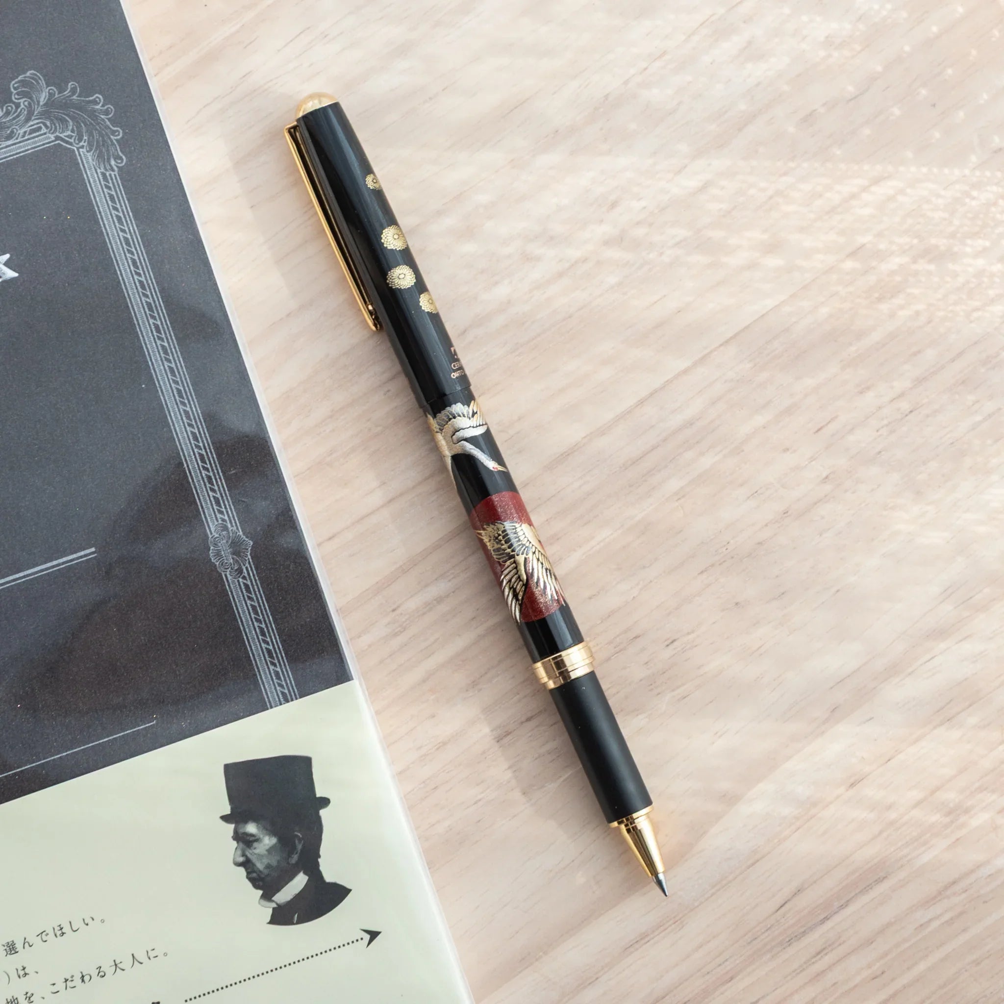 Overhead flat lay of black lacquer Japanese ballpoint pen with crane and sunrise pattern beside grey notebook on light wood