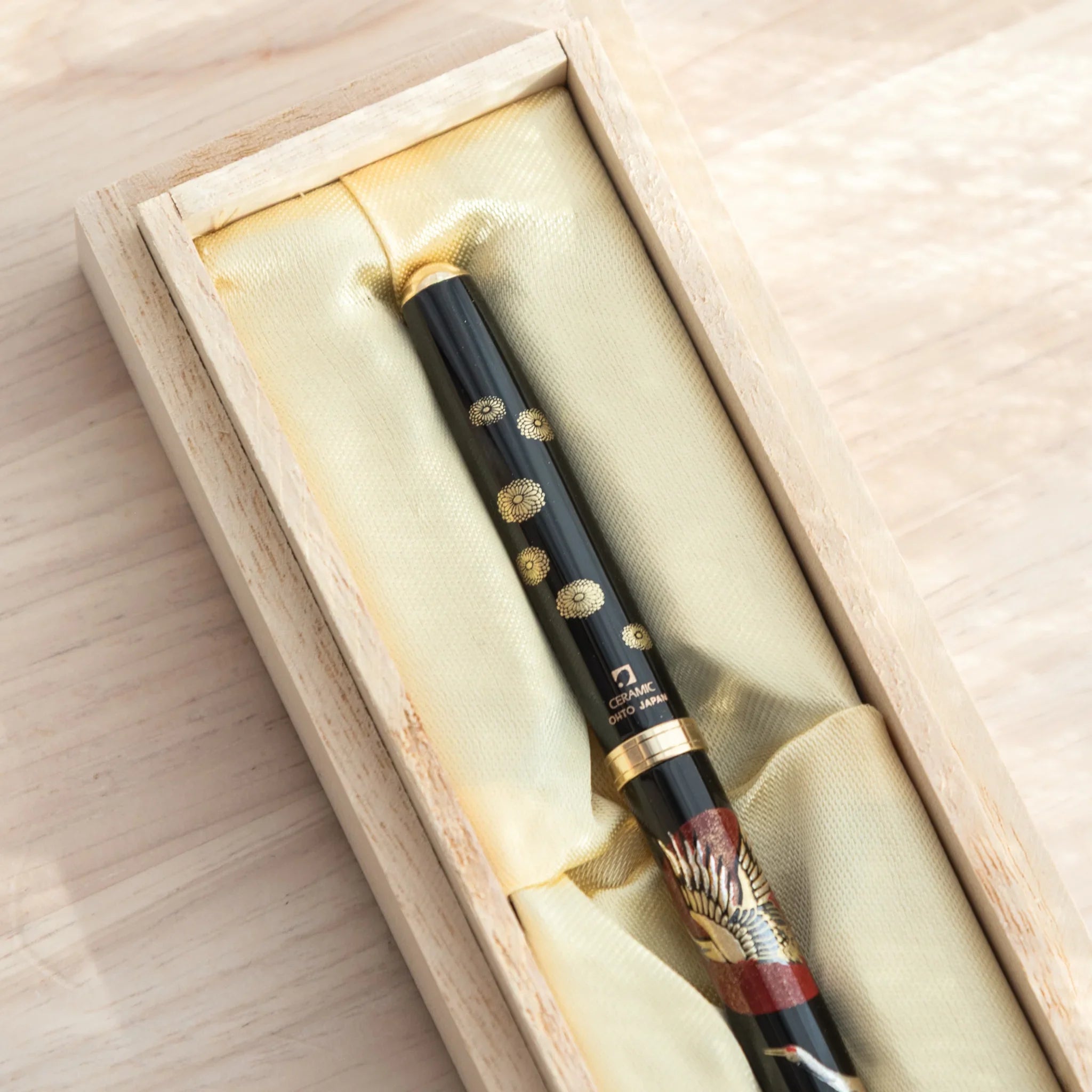 Cap end of Japanese ballpoint pen in open natural wood box showing gold chrysanthemum pattern on black barrel and cream satin