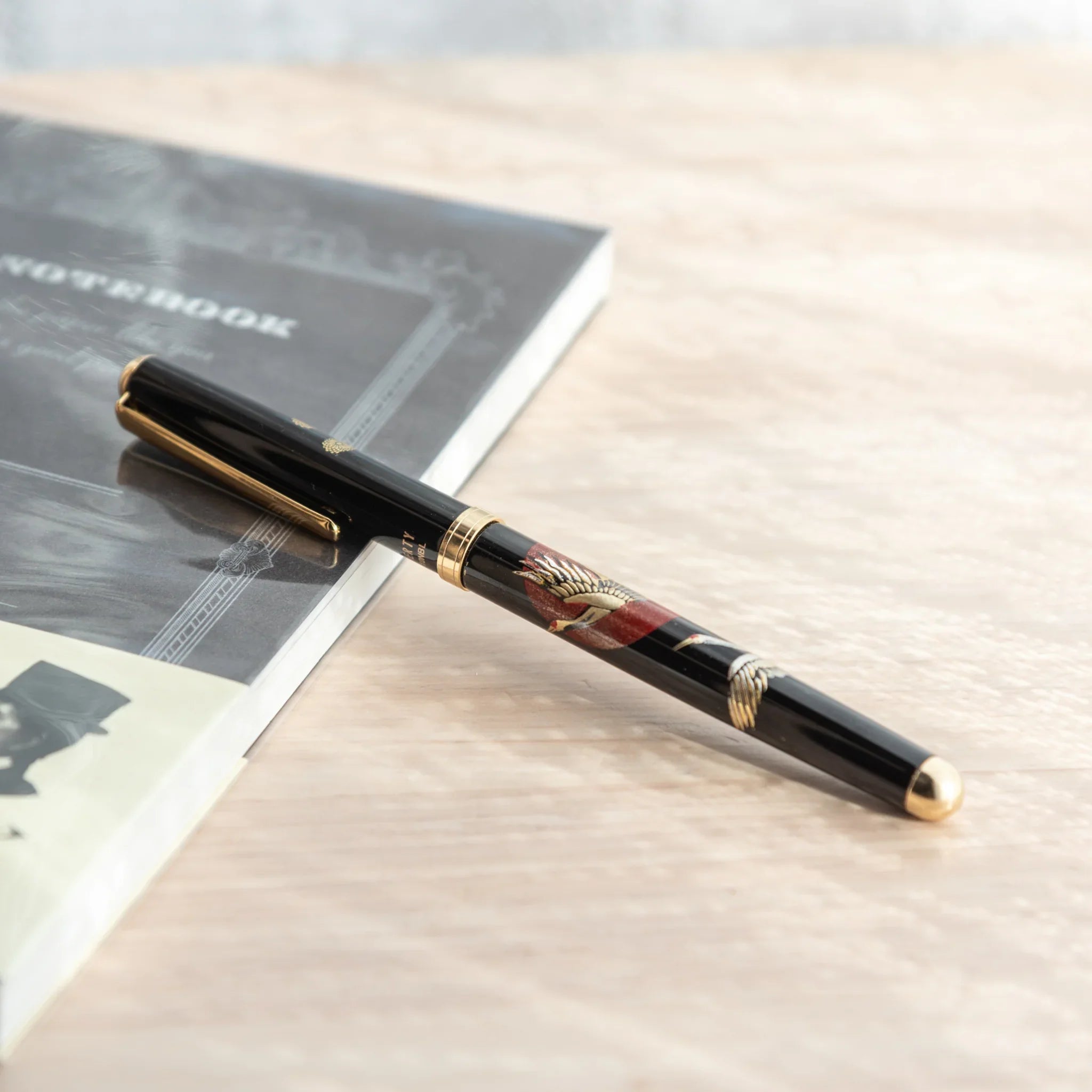 Black lacquer Japanese ballpoint pen with crane and sunrise pattern resting diagonally on grey notebook on light wood