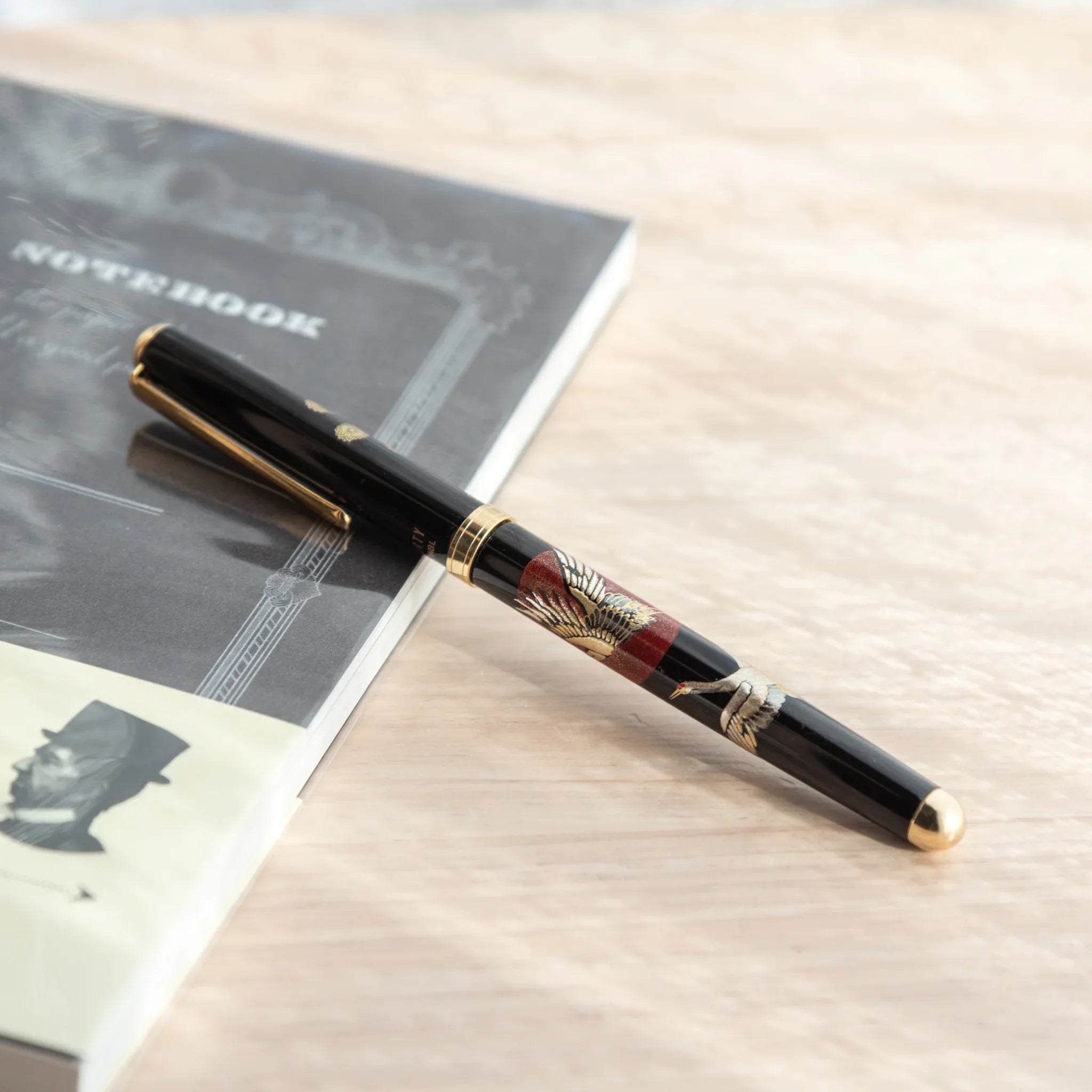 Black lacquer Japanese ballpoint pen with spread crane wings and red sunrise on barrel, angled on grey notebook