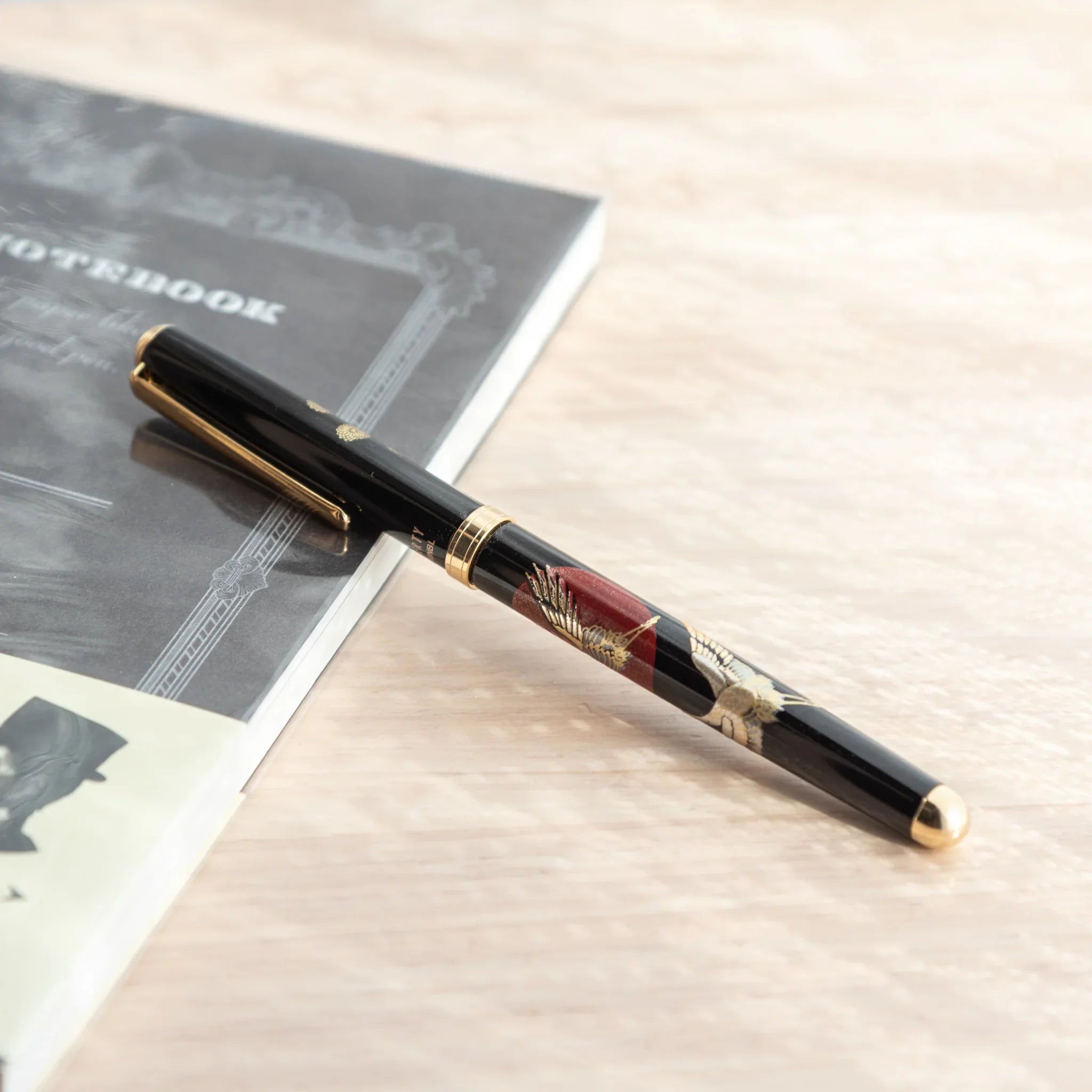 Black lacquer Japanese ballpoint pen with crane and sunrise barrel design at angle on grey notebook, gold end cap visible