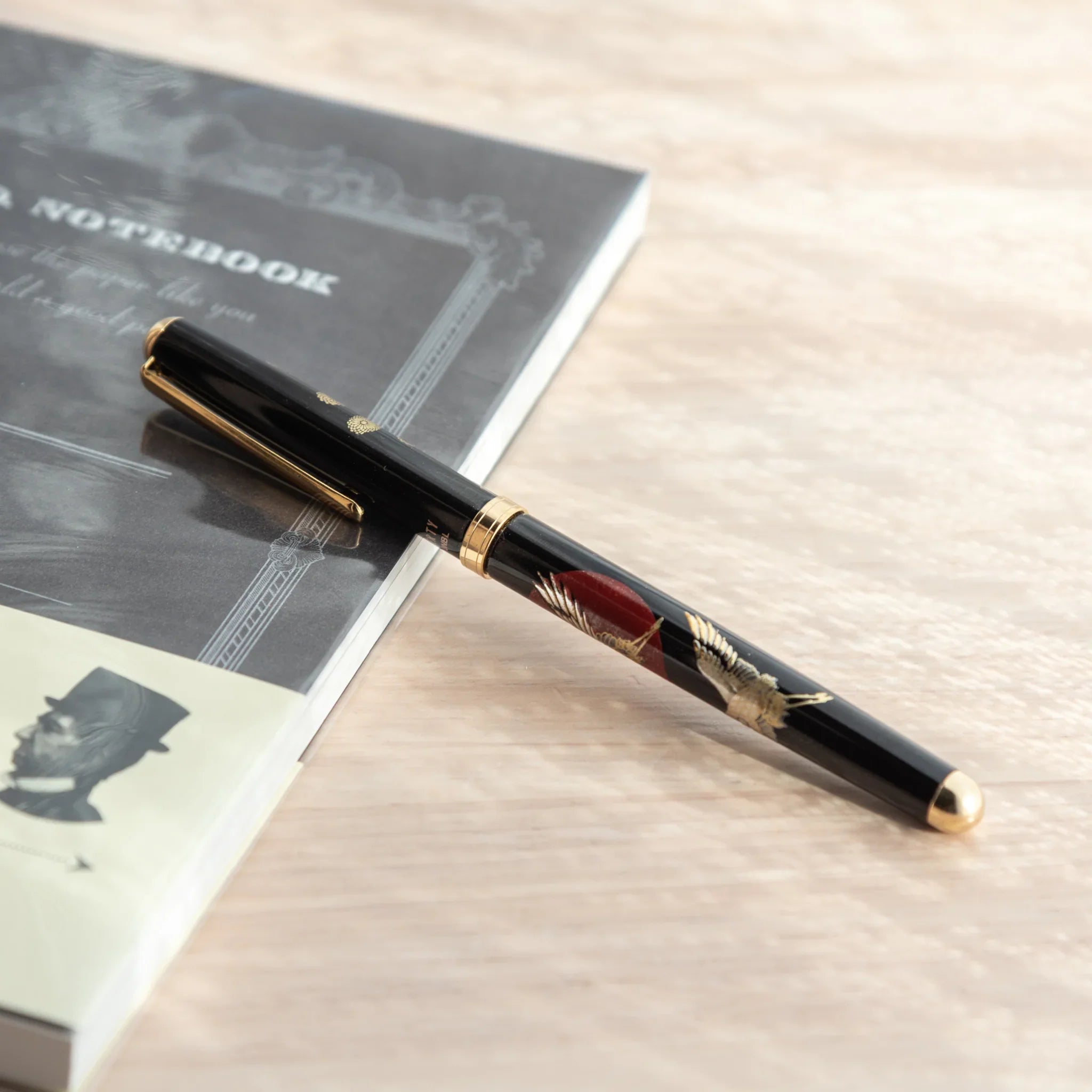 Black lacquer Japanese ballpoint pen with crane and sunrise pattern on grey notebook with Japanese text, viewed from above