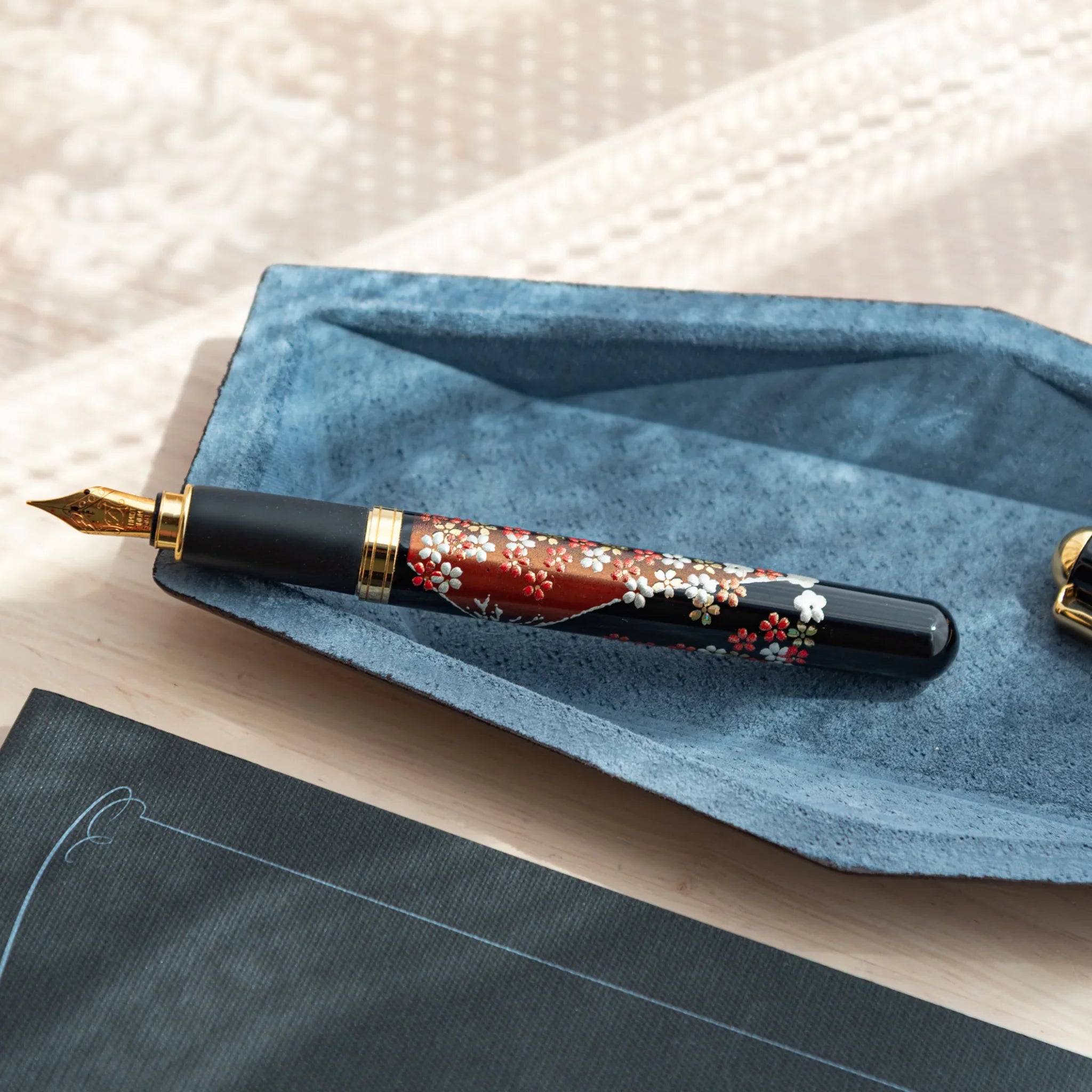 Uncapped Japanese fountain pen with red floral pattern and gold nib resting on a blue-gray suede hexagonal pen tray