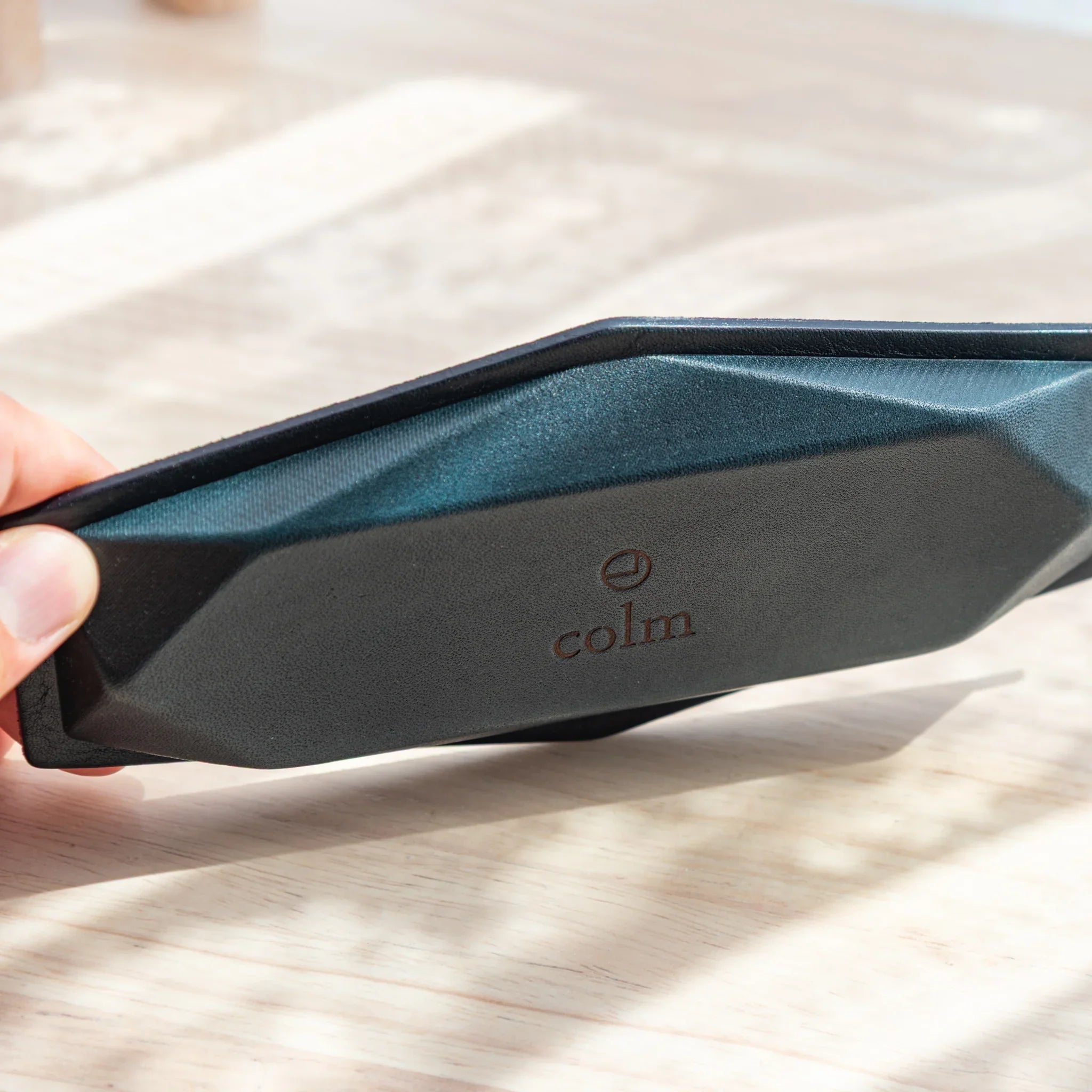 Hand tilting a blue-gray suede hexagonal slim pen tray to reveal the black leather base with embossed colm brand logo