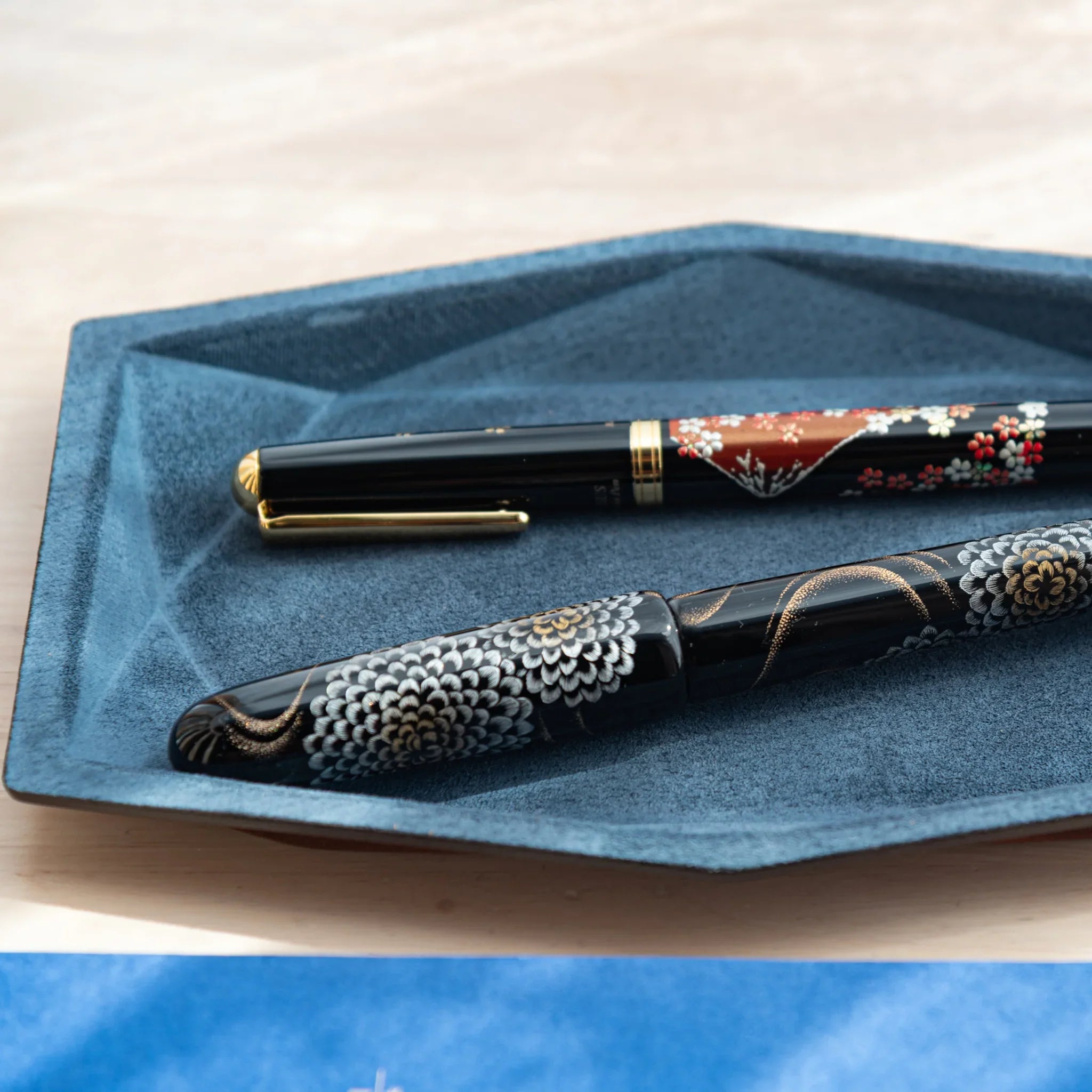 Blue-gray suede wide hexagonal pen tray from an angled view holding two Japanese fountain pens with floral patterns