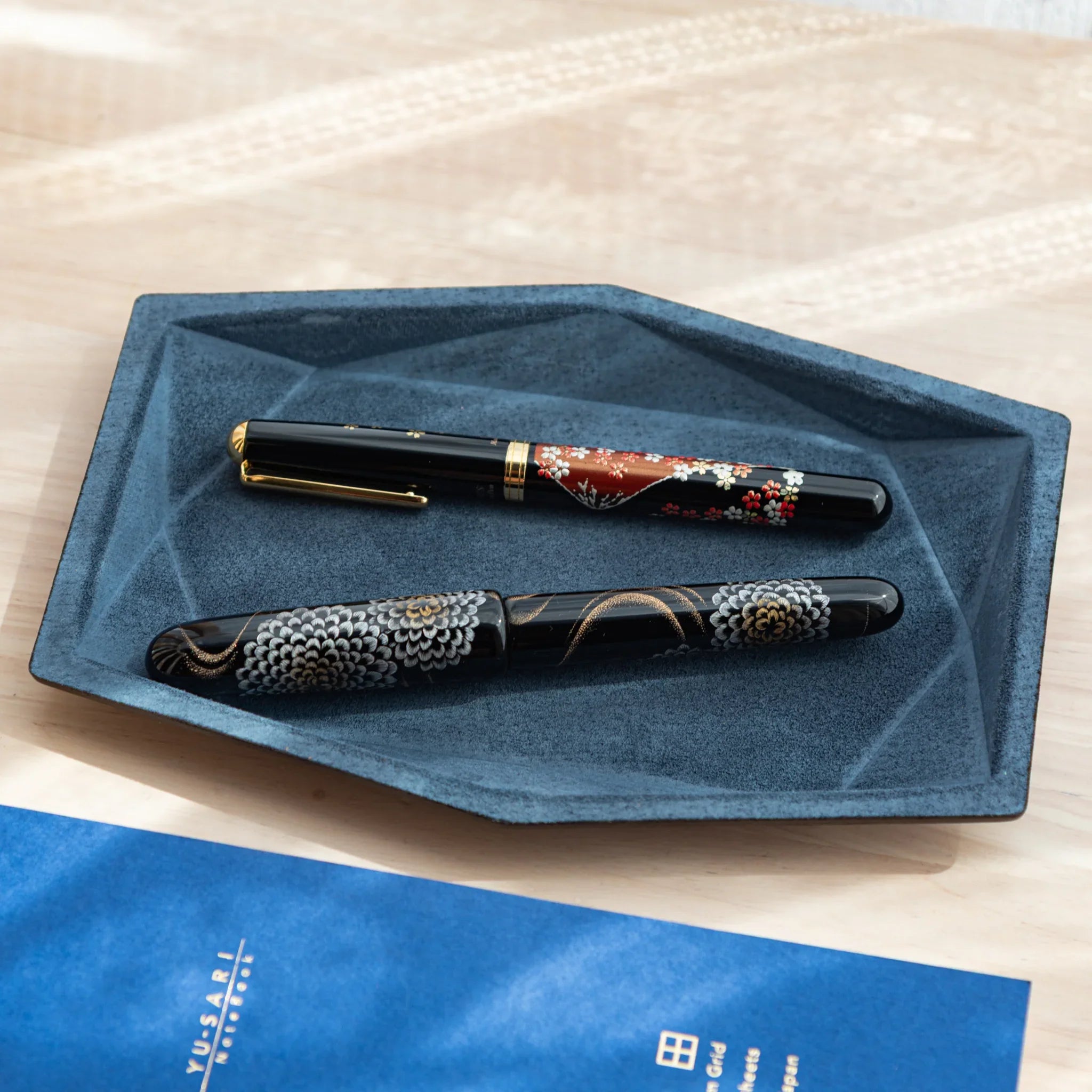 Blue-gray suede wide hexagonal pen tray holding two Japanese fountain pens with sakura and chrysanthemum patterns overhead