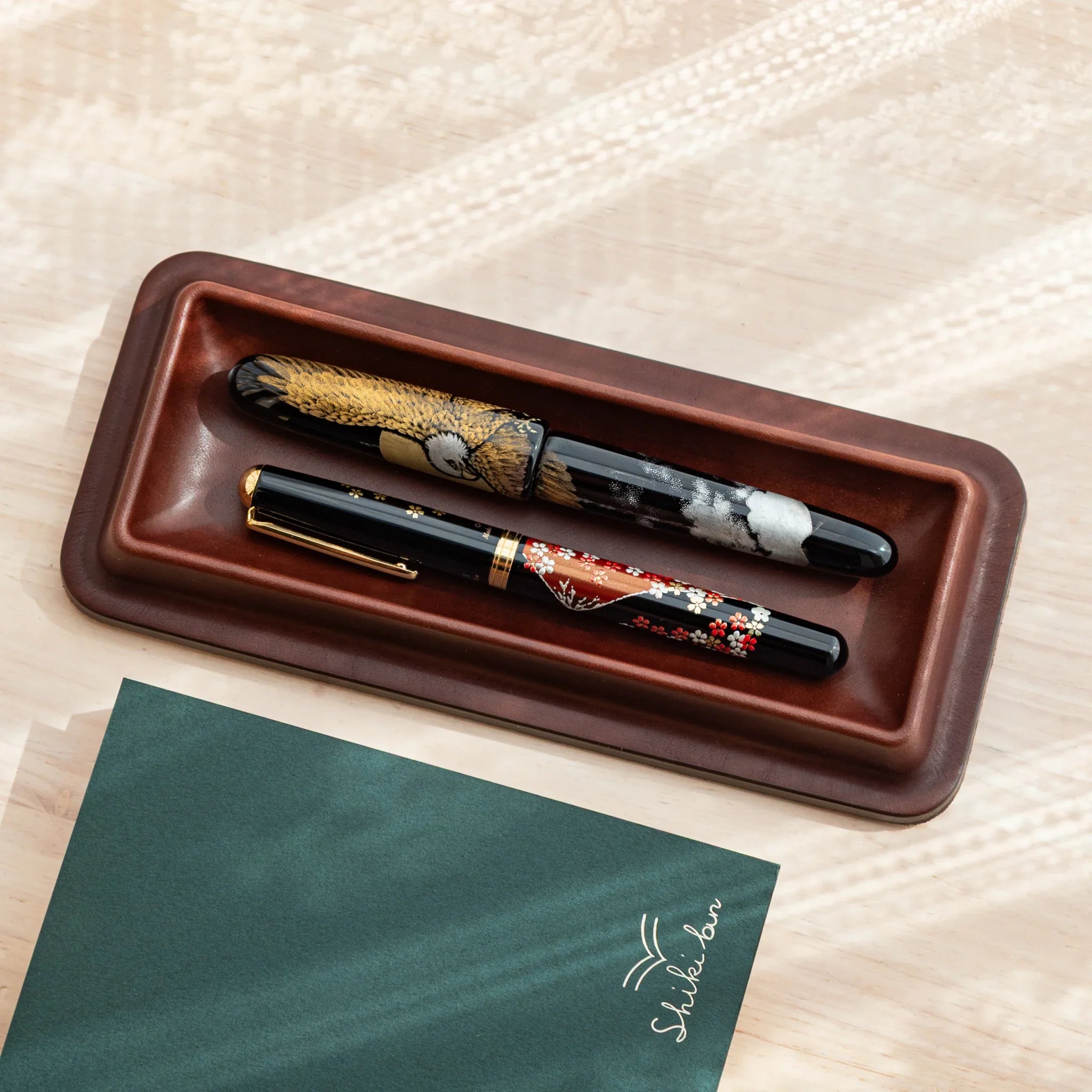 Burgundy leather pen tray displaying multiple decorative pens including gold trimmed and ornate models