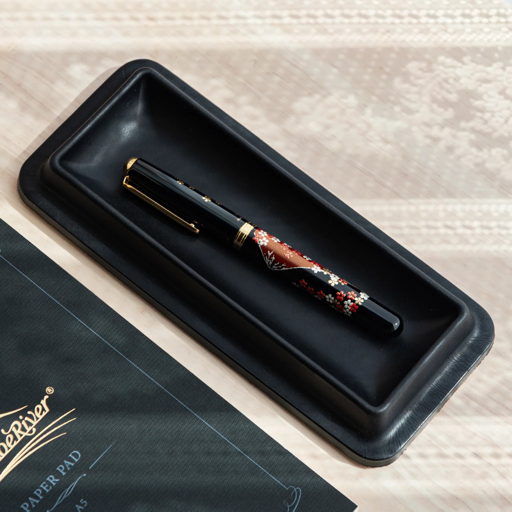Black leather pen tray containing red and burgundy decorative maki-e pen with gold trim and attached