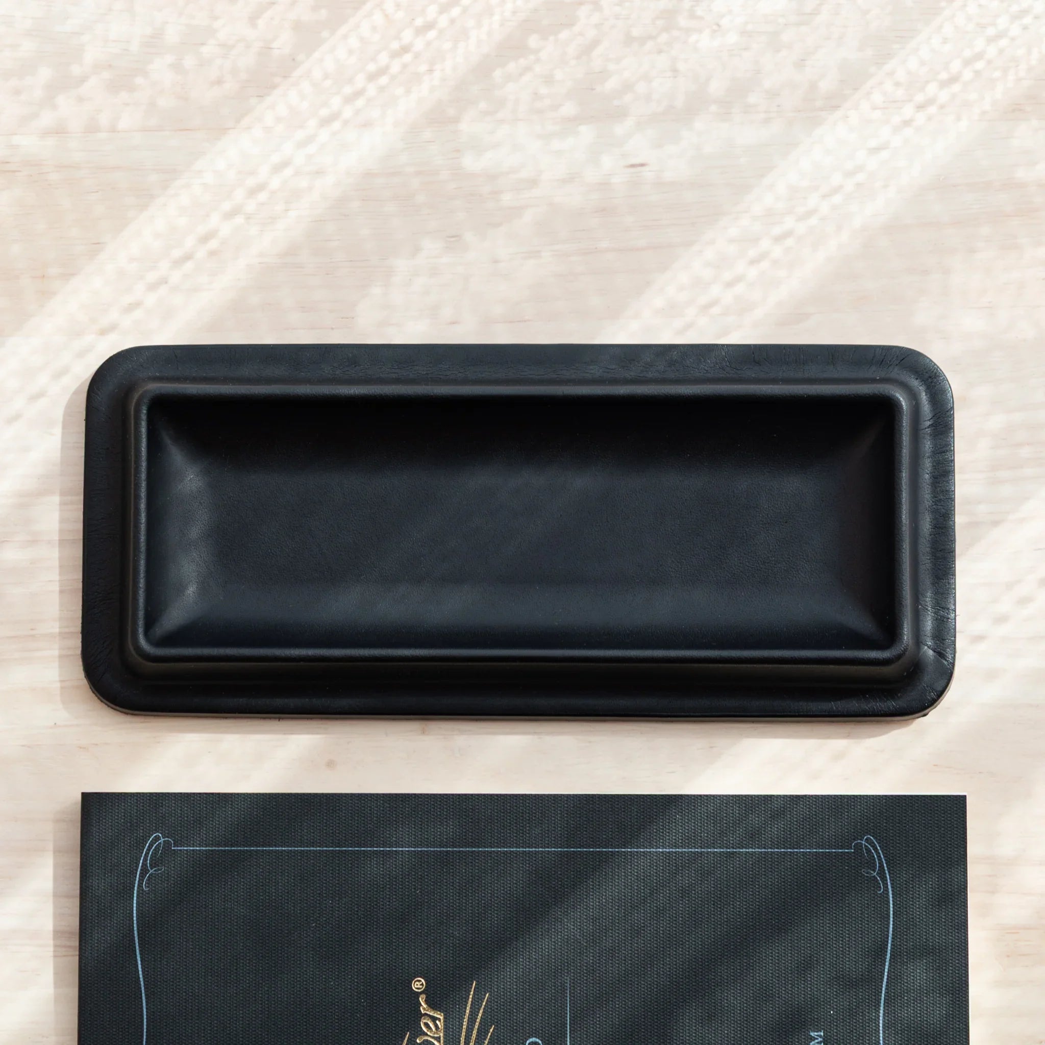 Empty black leather pen tray shown from directly overhead with dark writing paper pad positioned below