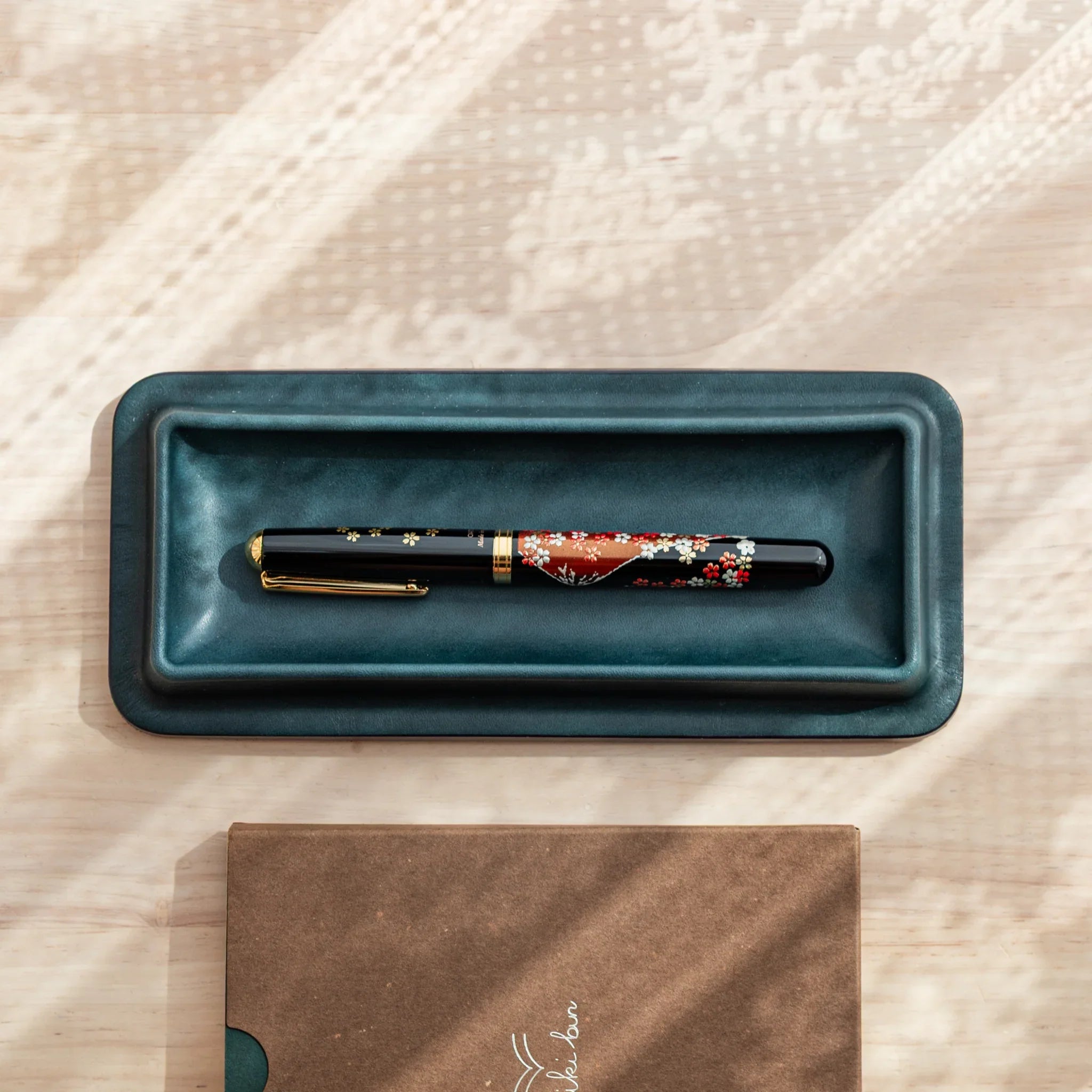 Olive green leather pen tray angled showing red patterned decorative maki-e pen inside the holder tray