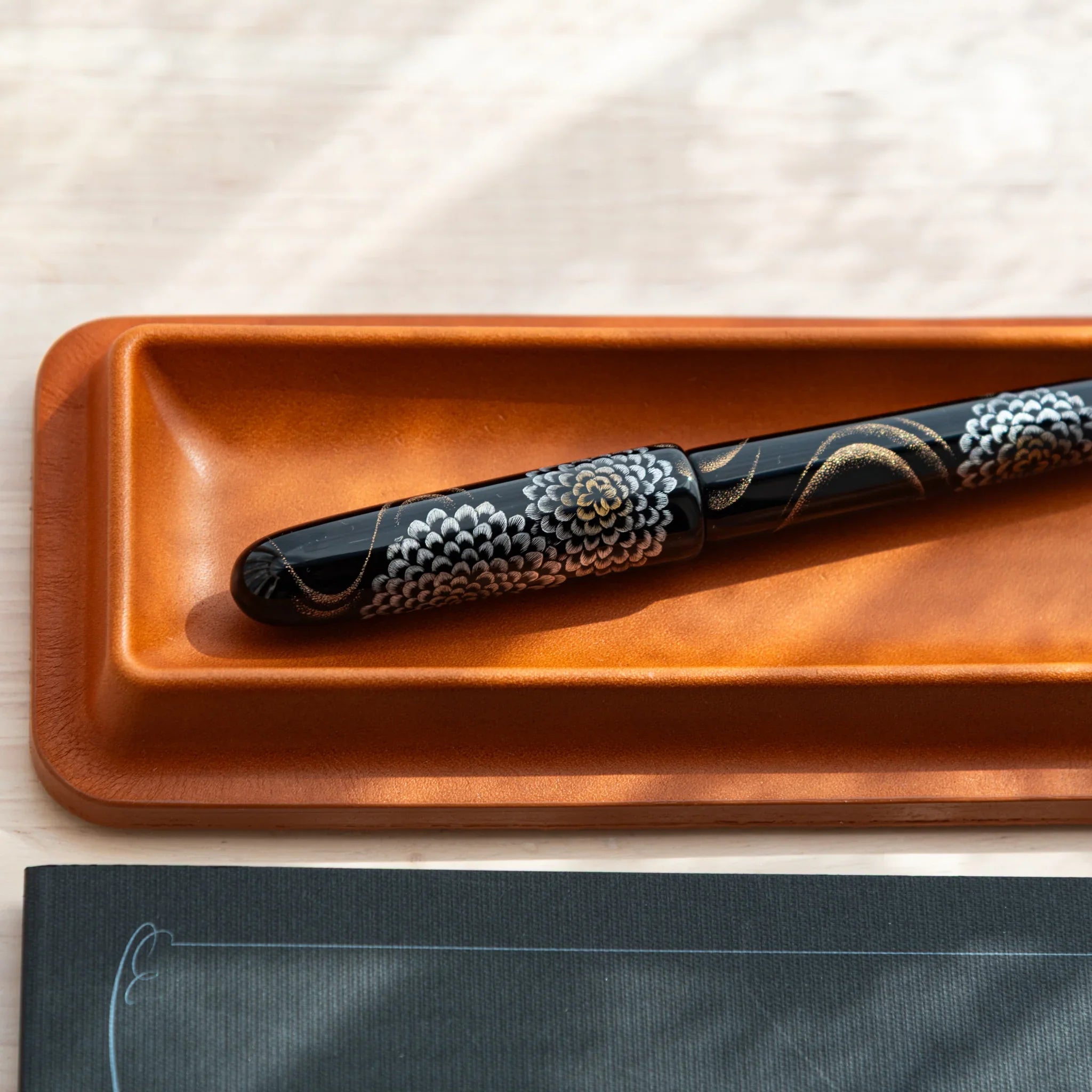 Cognac colored leather pen tray angled view displaying black decorative maki-e pen with white dot pattern