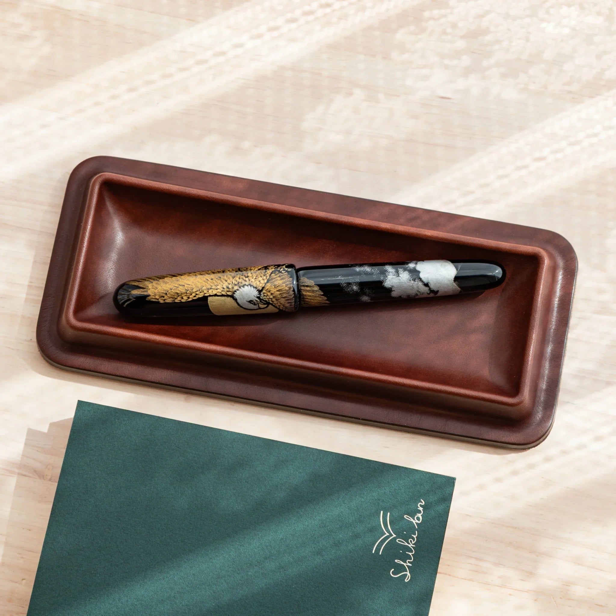 Dark burgundy leather pen tray overhead containing brown decorative maki-e pen and writing paper pad