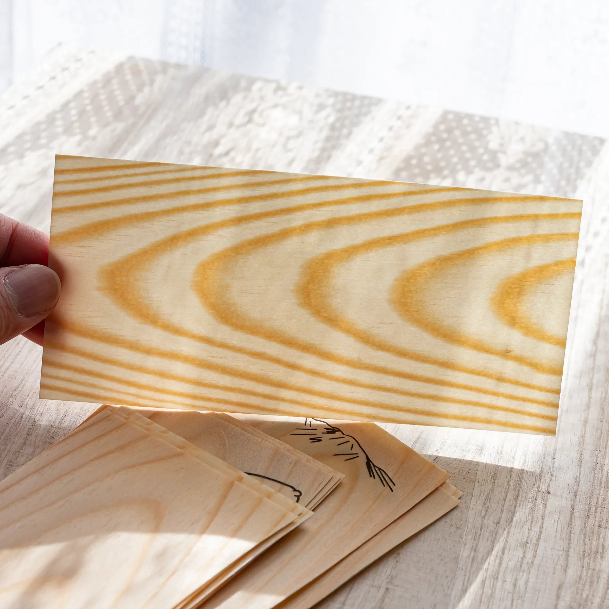Japanese Stationery thin wood message card held up to show its natural pine grain, with stacked cards below on a desk