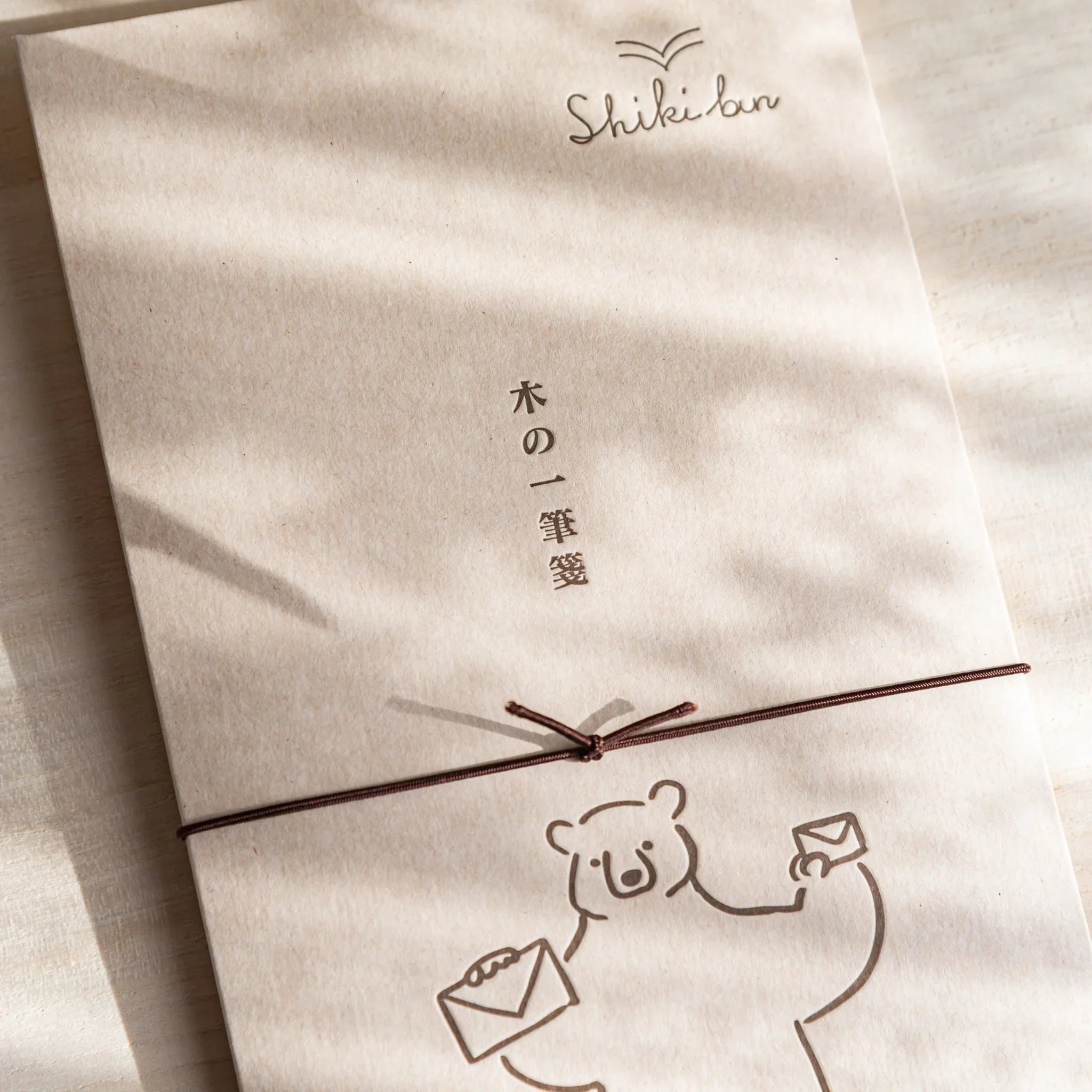 Thin wood message card wrapped with brown string, featuring a bear illustration and soft curtain shadows on a light desk