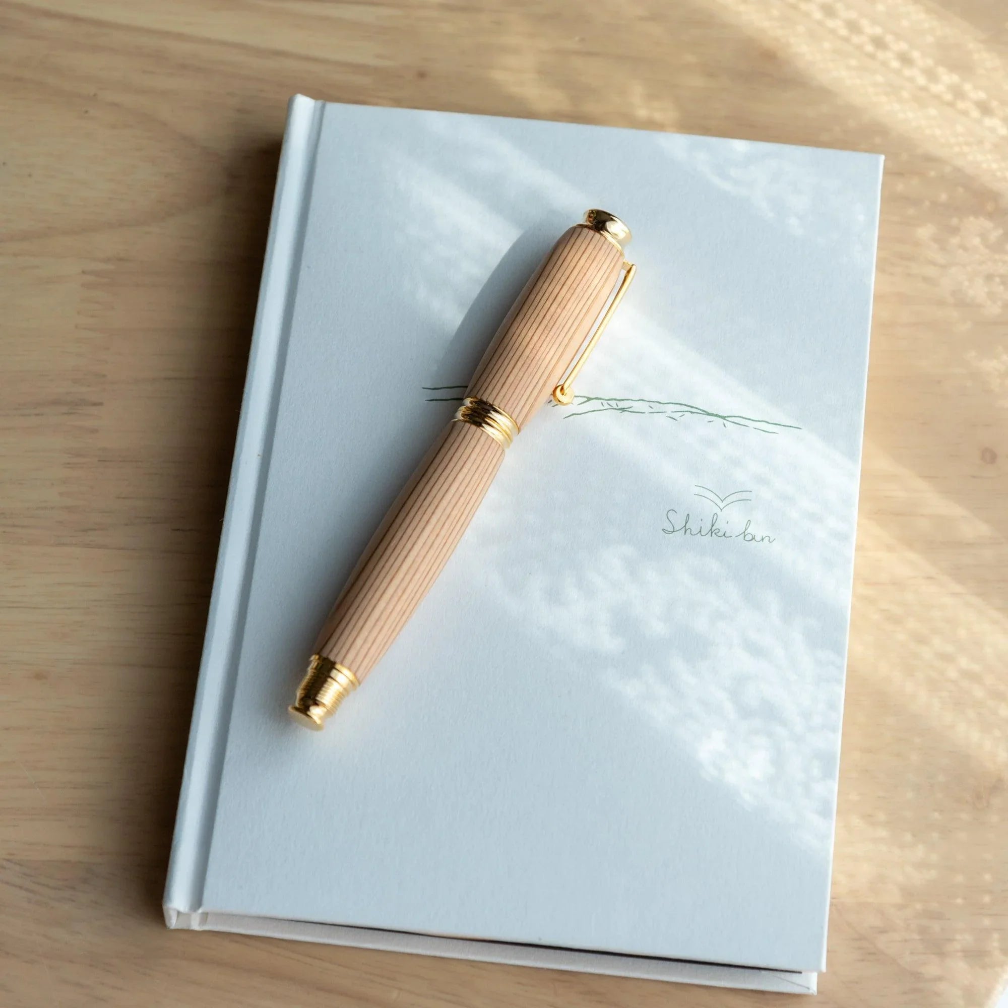 Pale cedar wood Japanese fountain pen body resting on a light blue Shiki notebook on a wood desk, gold trim rings visible