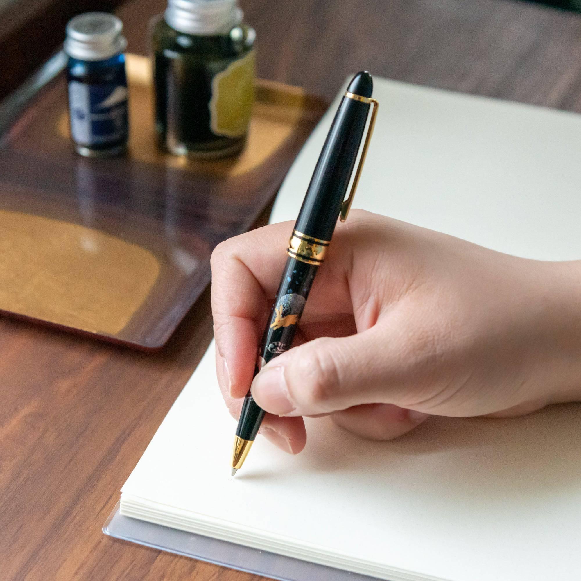 Japanese ballpoint pen black lacquer barrel with gold makie rabbit moon design, held over paper with ink bottles