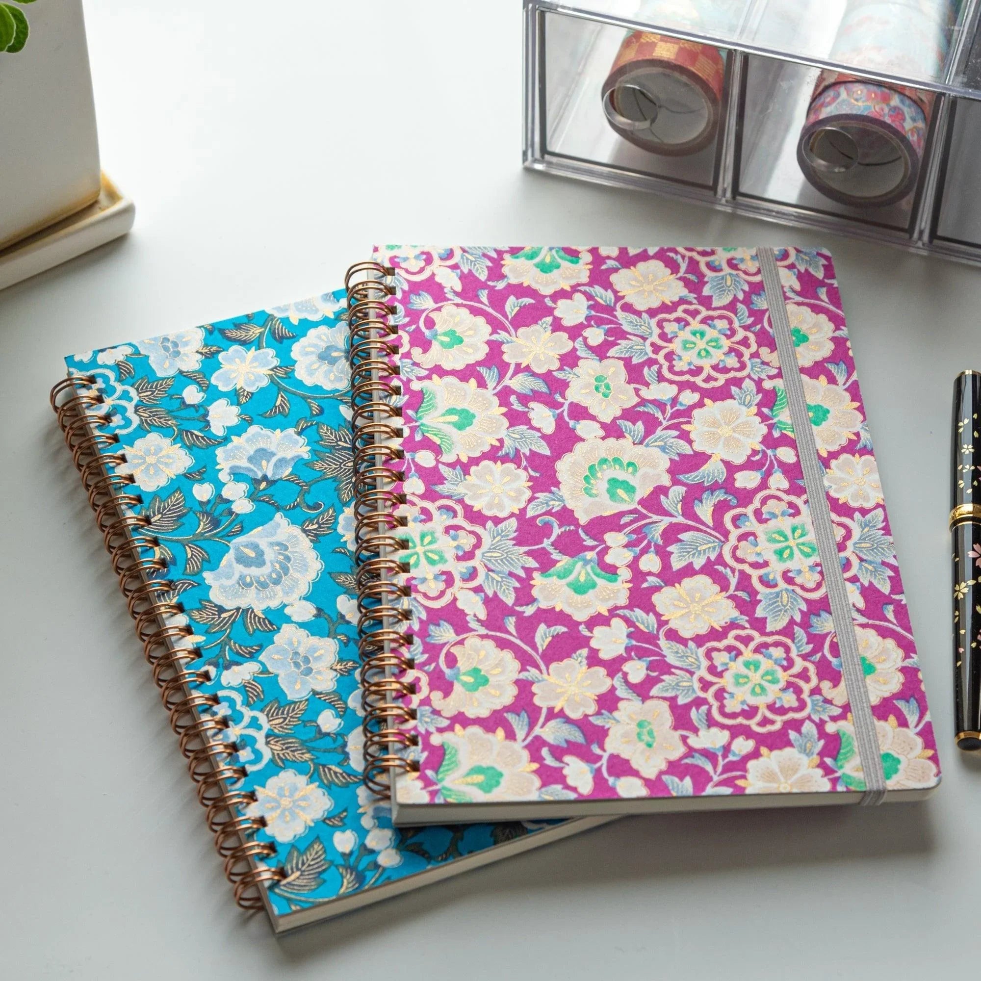 Two yuzen washi Japanese notebooks in magenta and teal on a desk at an angle, with a fountain pen and washi tape rolls nearby